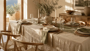 A rustic Mediterranean-style dining table set with an oatmeal linen tablecloth, sage green runner, and a central olive branch arrangement in a stone vase.