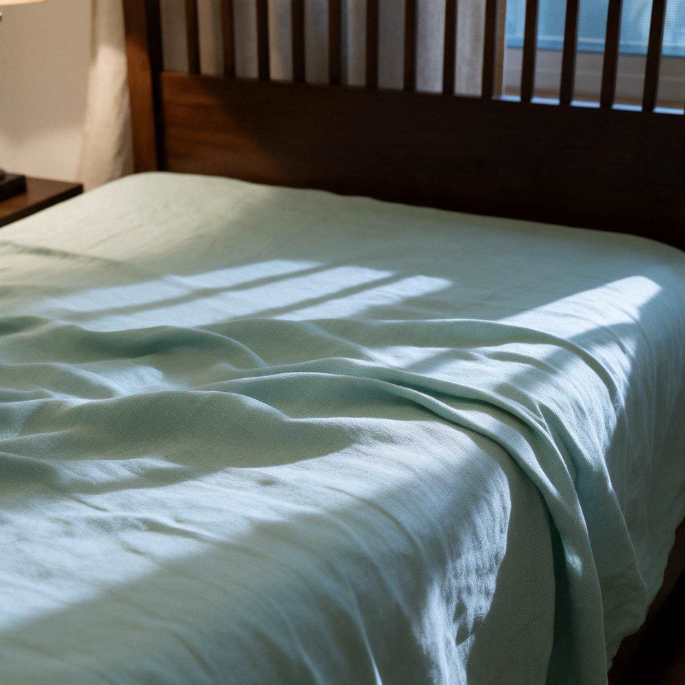 Blue Surf linen flat sheet and matching pillowcases on a dark wood slatted bed, styled with a classic table lamp and red clock on the bedside table in warm ambient light
