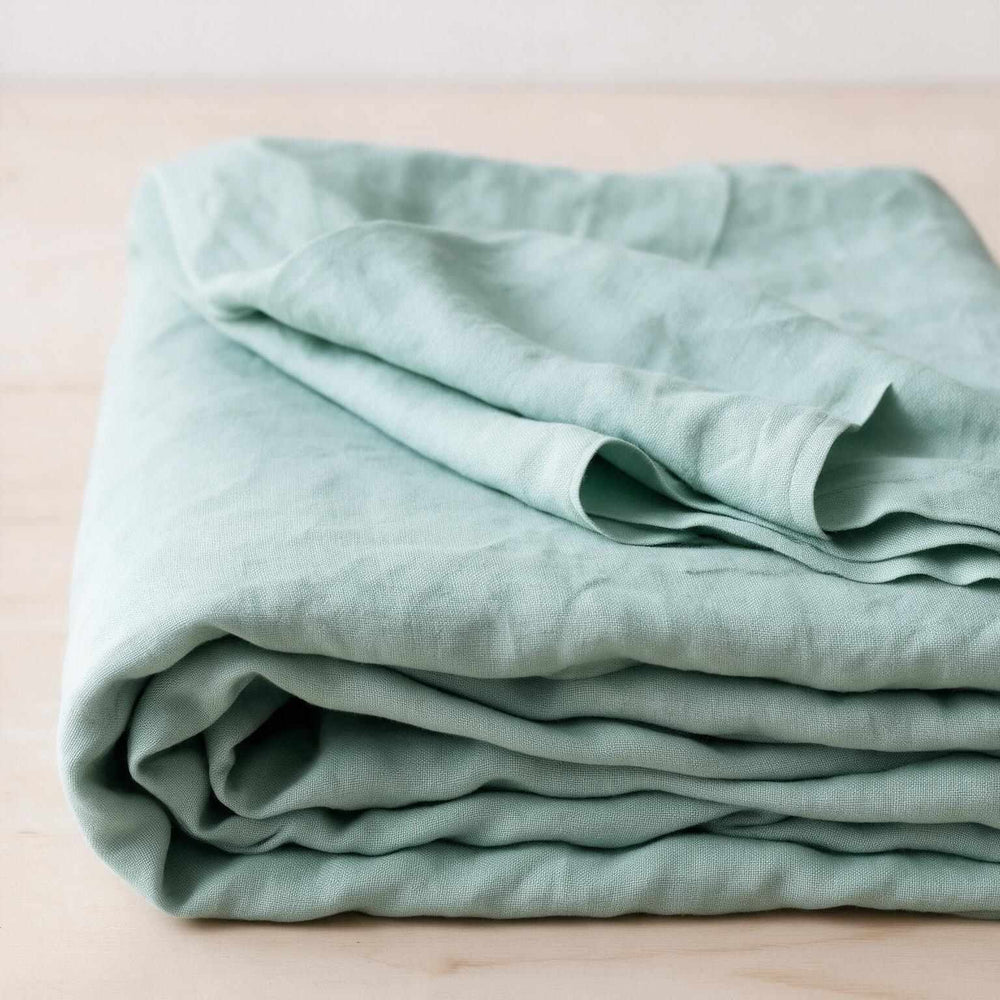Blue Surf linen flat sheet loosely draped across a bed with soft window light casting geometric shadows, highlighting the natural drape and stonewashed sage-blue linen texture