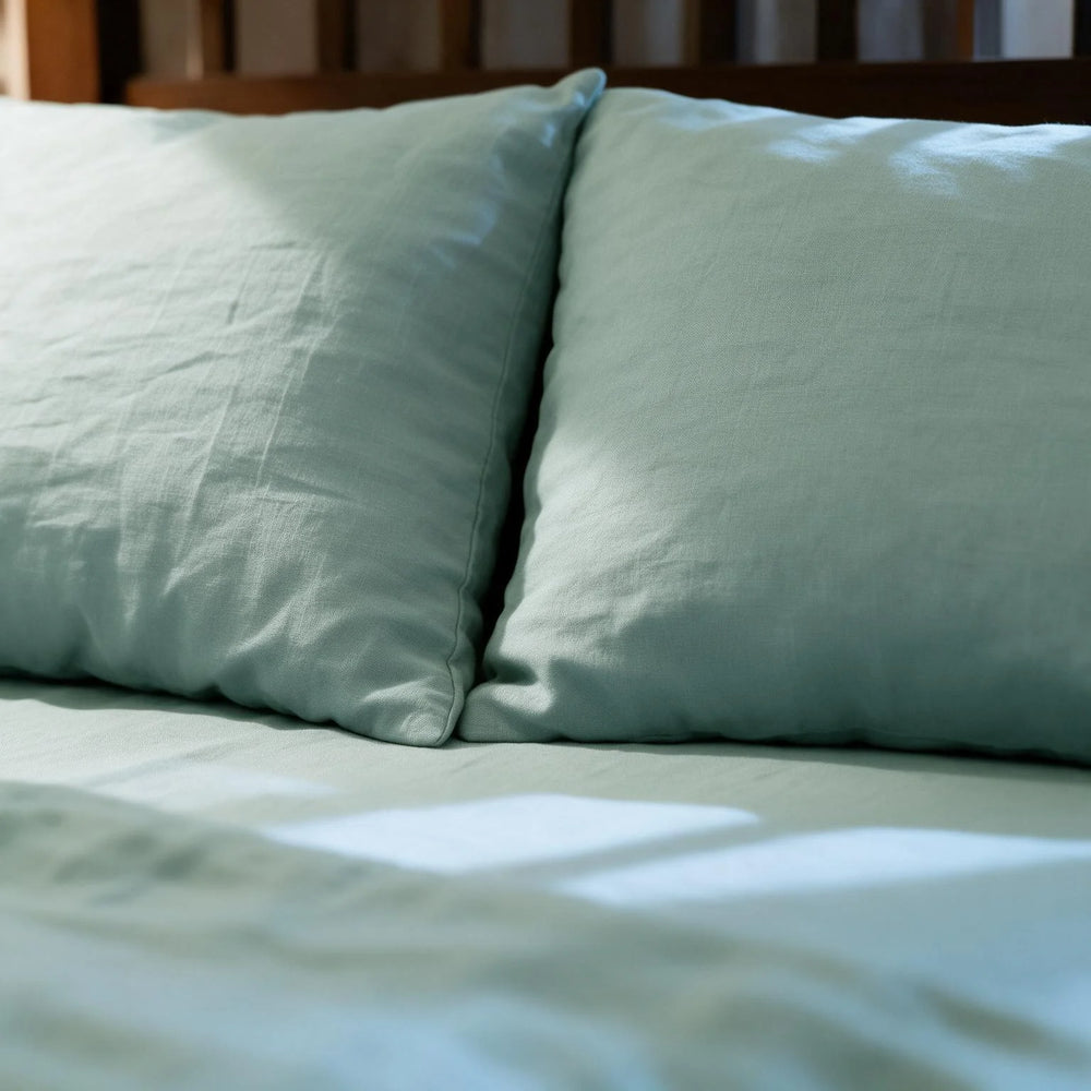 Two Blue Surf linen pillowcases resting against a wooden headboard with a matching flat sheet beneath, sage-blue stonewashed linen in warm natural light