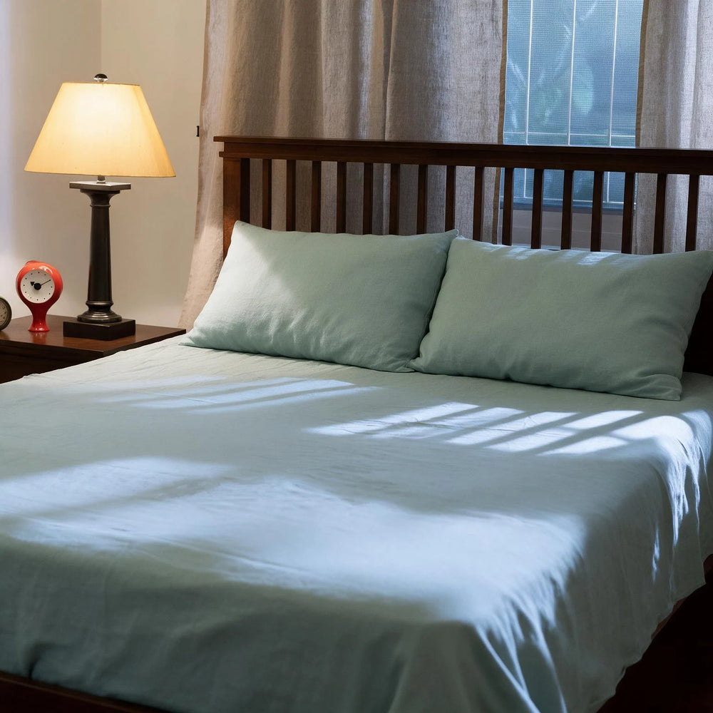 Blue Surf linen flat sheet set styled on a full bed with matching pillowcases, dark wood headboard, bedside lamp, and sheer curtains in a warm bedroom setting
