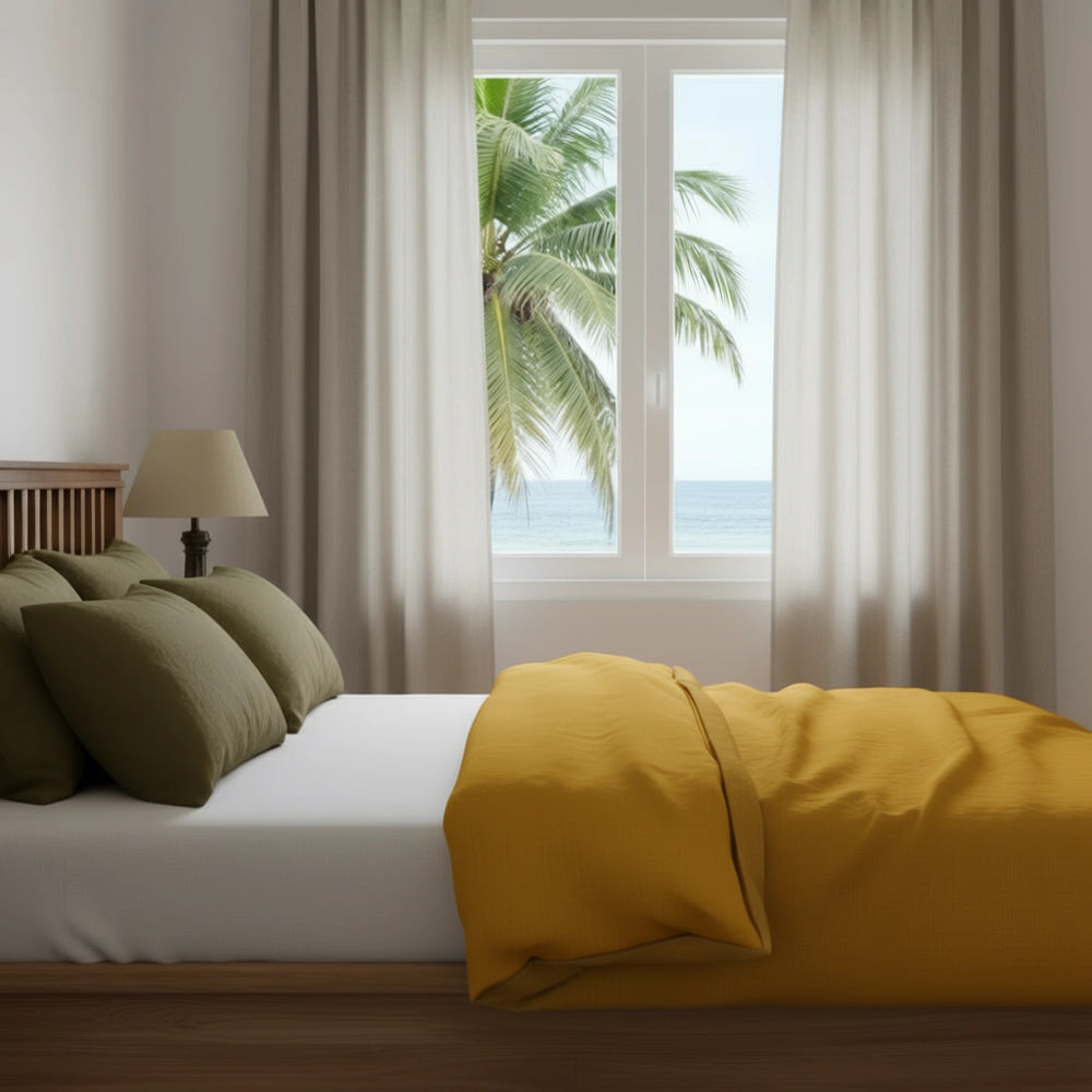 Bedroom with a bed featuring yellow bedding and green pillows, large window with white curtains, and view of palm trees and ocean.