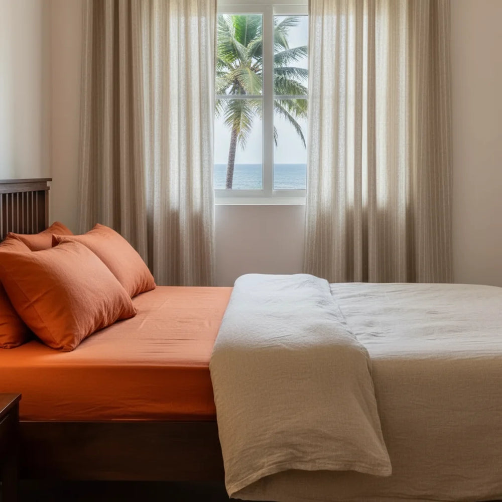 Bed with orange bedding and beige blanket in a room with a window showing palm trees and ocean.