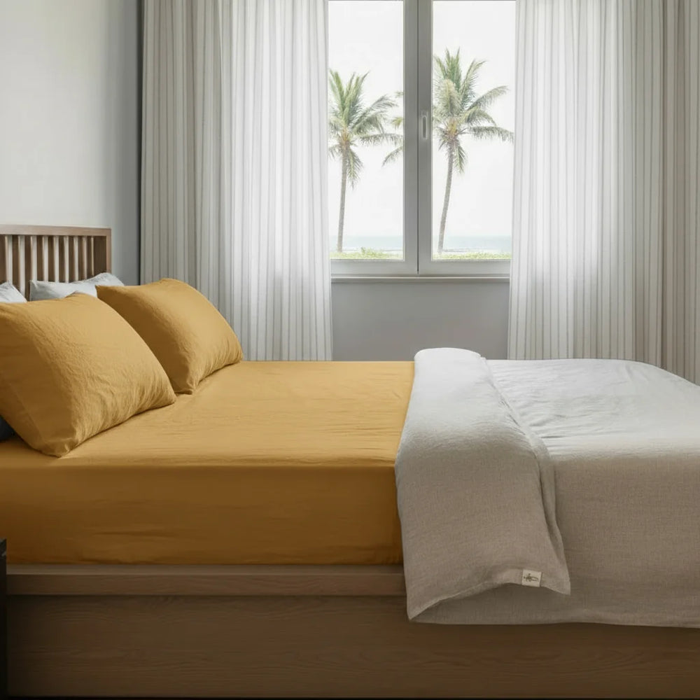 Bed with yellow bedding and white comforter in a room with large windows and palm trees outside.