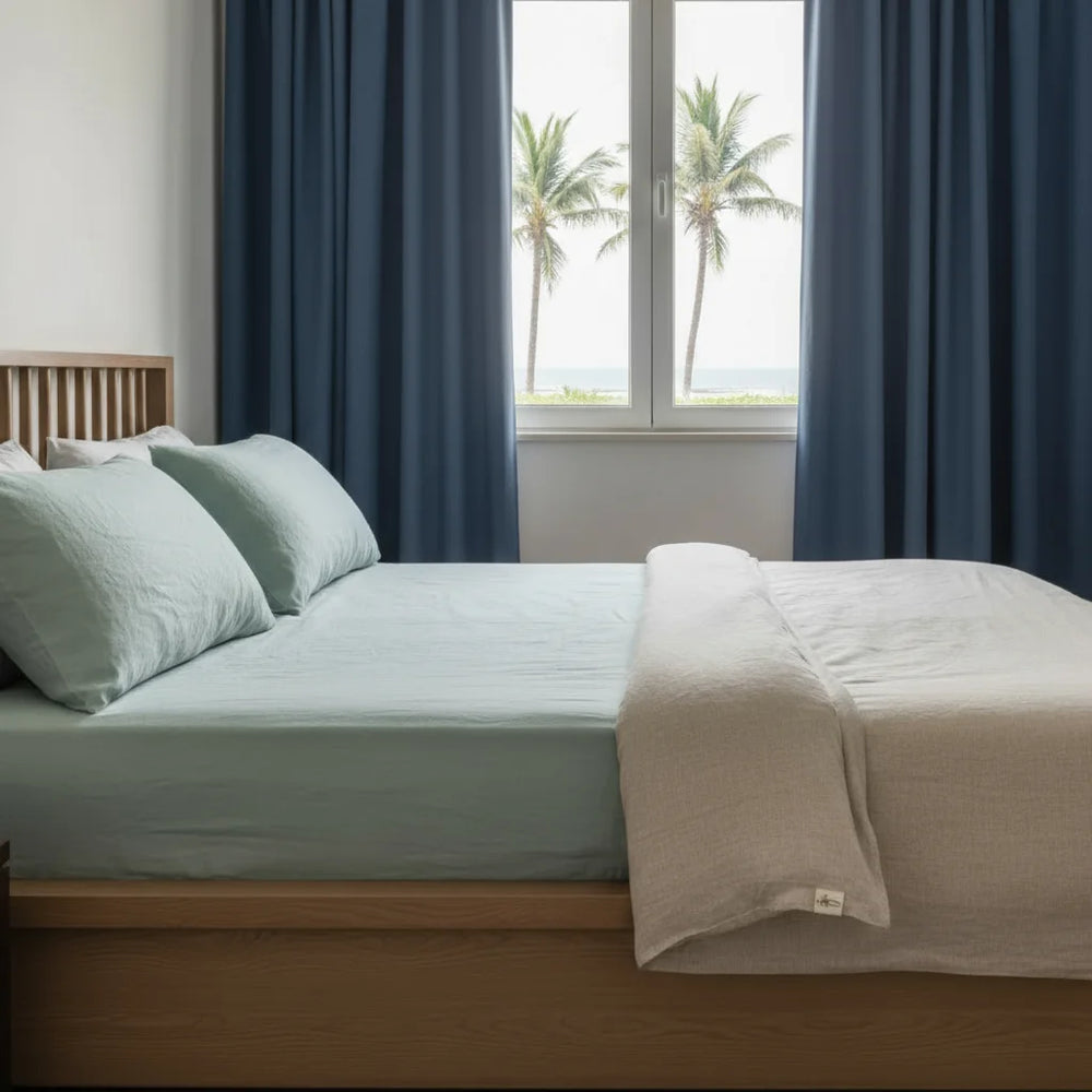 Bedroom with a bed featuring light blue bedding and a beige blanket, large windows with blue curtains, and palm trees outside.