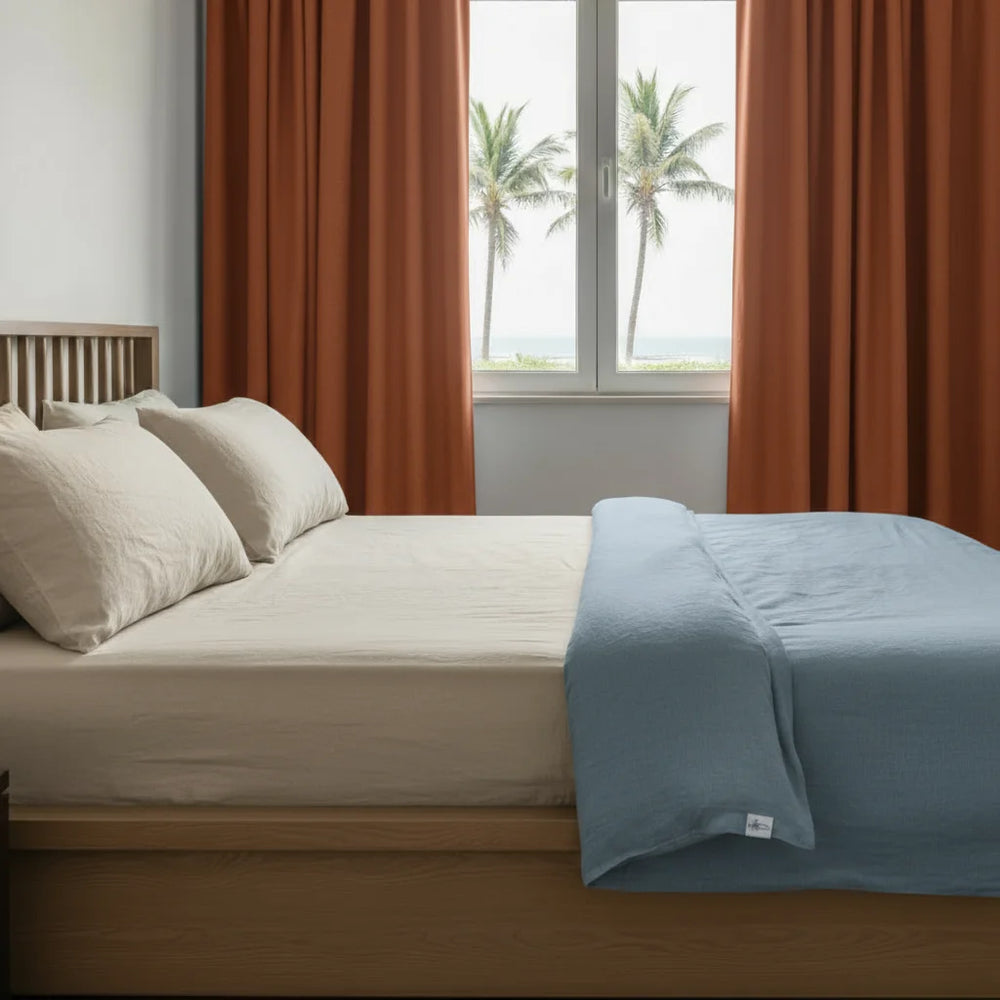 Bedroom with a bed featuring white bedding and a blue blanket, with orange curtains and a window view of palm trees.