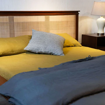 A bed dressed in mustard yellow bedding with a textured light gray pillow, set against a woven headboard, with a dark blue blanket draped in the foreground and a bedside lamp on a nightstand.