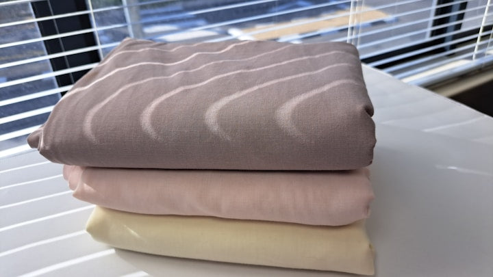 Three folded fabric pieces stacked on a white table, with a window and blinds in the background. The top fabric is taupe with a wavy pattern, the middle is light pink, and the bottom is cream.
