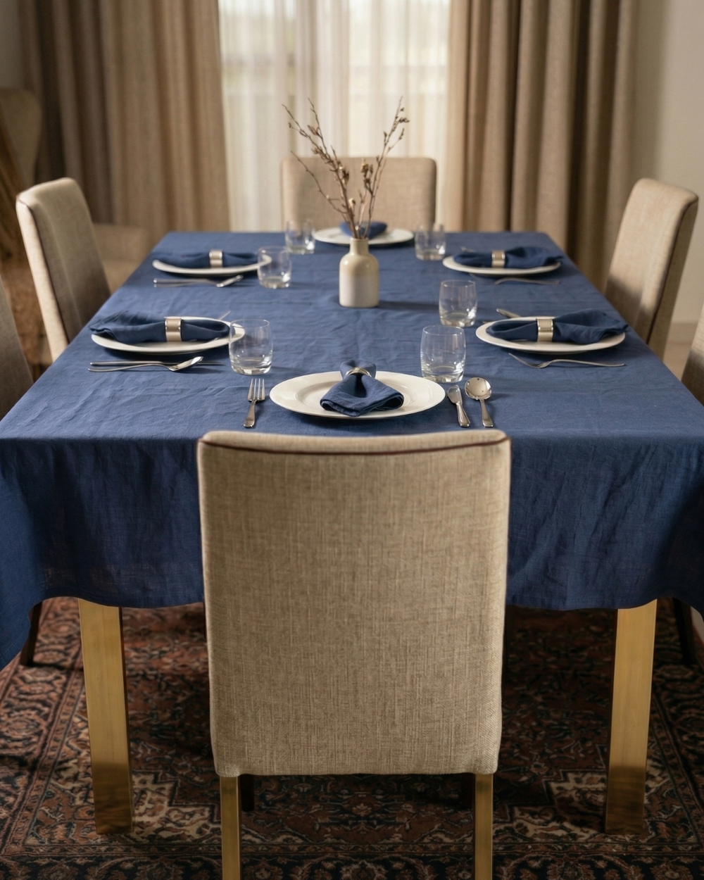 Dining table set with blue tablecloth, plates, and chairs in a room with curtains.