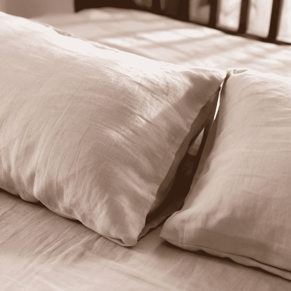 Two Natural Beige linen pillowcases resting on a matching flat sheet against a dark wood headboard, soft warm light highlighting the relaxed linen texture