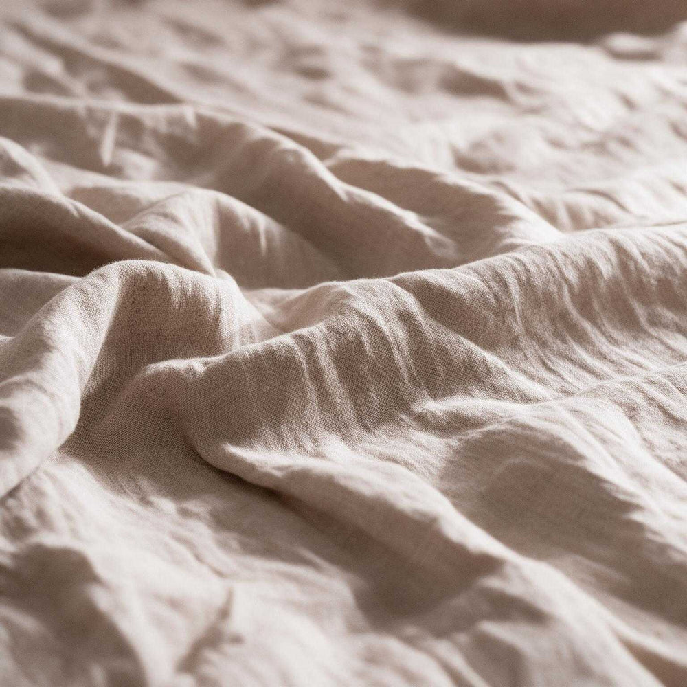 Two Natural Beige linen pillowcases against a dark wood slatted headboard with a classic table lamp and clock on the bedside table, warm light filtering through sheer curtains⠋
