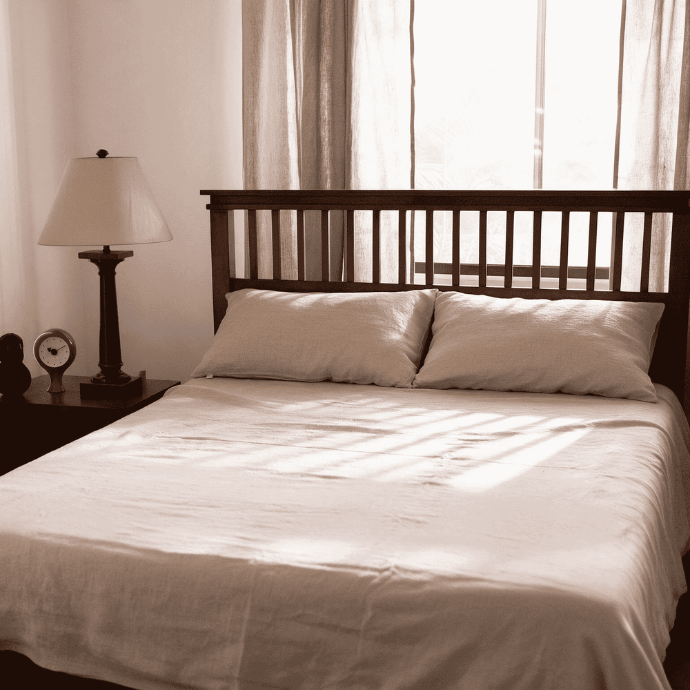 Natural Beige linen flat sheet set styled on a full bed with matching pillowcases, dark wood headboard, bedside lamp, and sheer curtains with soft morning light
