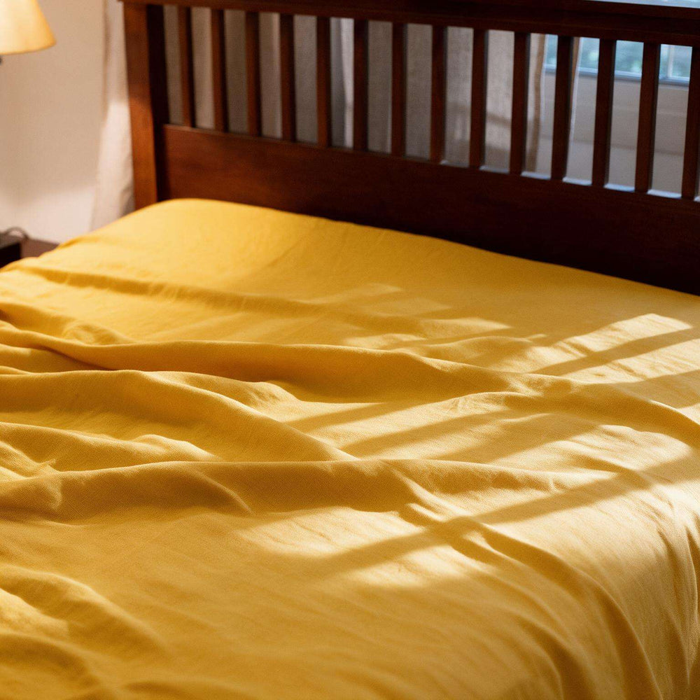Nugget Gold linen flat sheet loosely draped on a dark wood slatted bed with warm window light casting soft shadows across the rich golden stonewashed linen