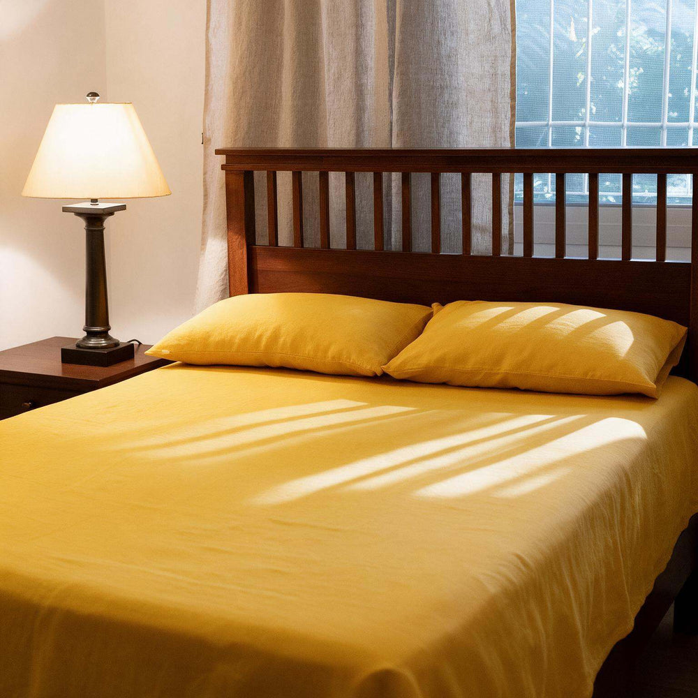 A neatly made bed with bright yellow bedding and two matching yellow pillows in a bedroom setting, with a wooden headboard, bedside lamp, curtains, and window visible.