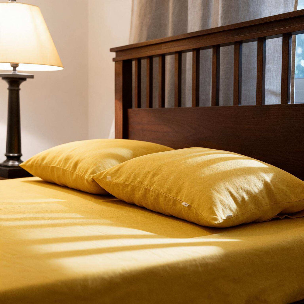 A bed dressed in a mustard yellow sheet and two matching yellow pillows, styled in a bedroom with a dark wood headboard, a lit table lamp, and soft window light.