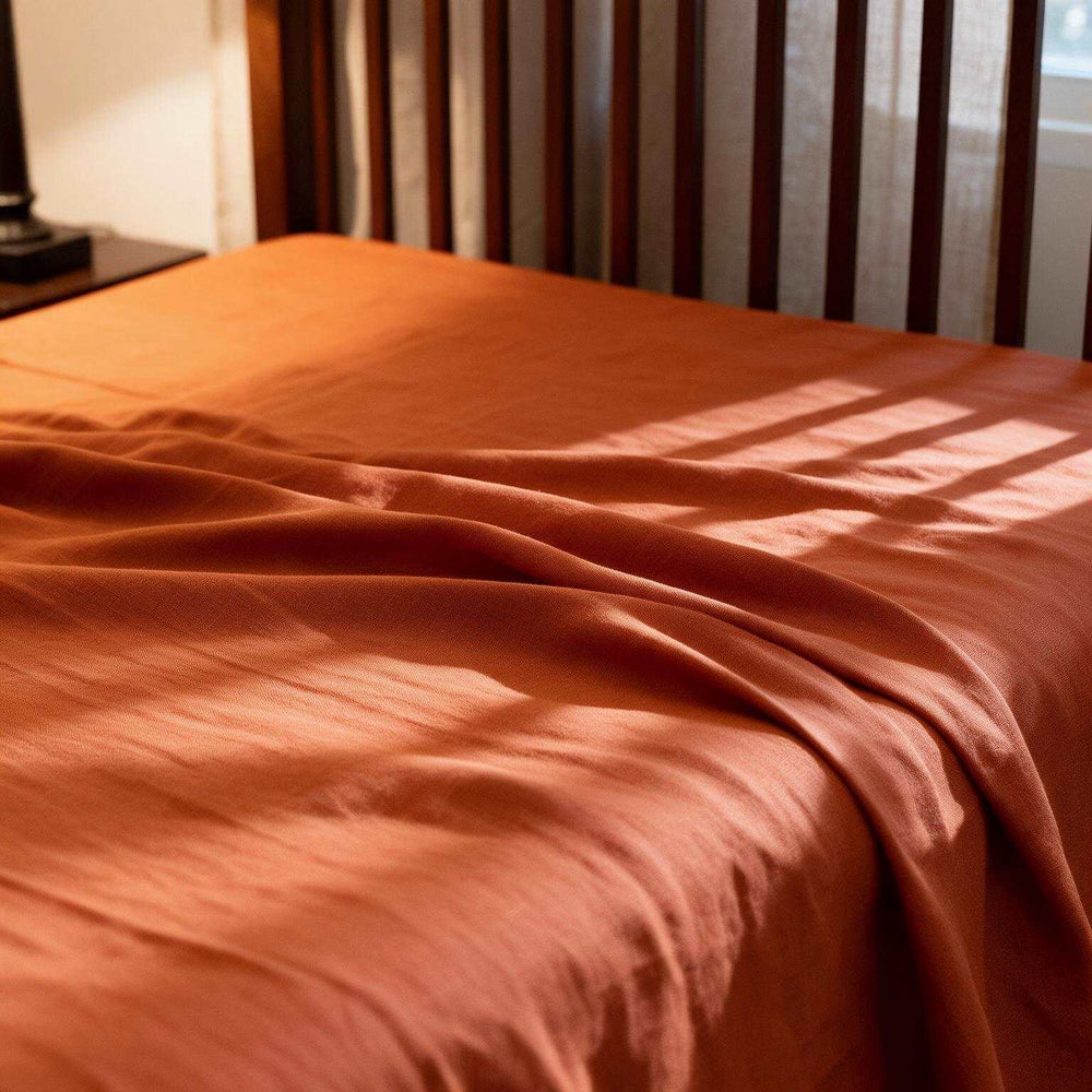 Rust Orange linen flat sheet and matching pillowcase on a dark wood slatted bed, styled with a classic table lamp and red clock on the bedside table in warm amber light