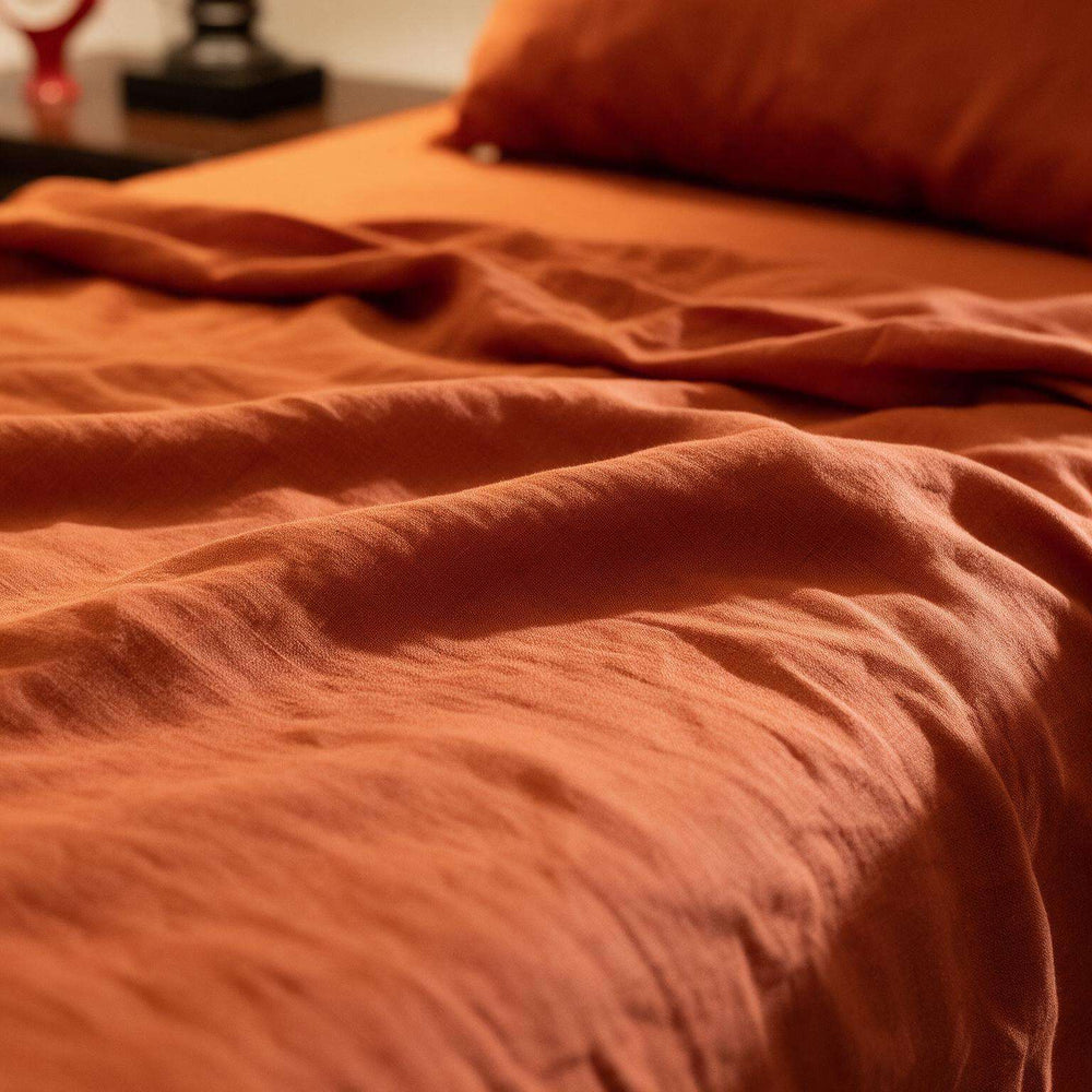 Close-up of two Rust Orange linen pillowcases on a matching flat sheet against a dark wood headboard, highlighting the rich terracotta stonewashed linen texture in warm light