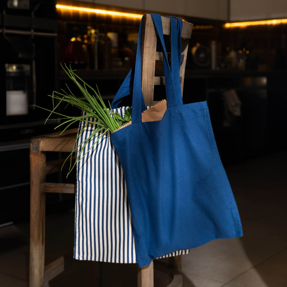 Two cotton shopping bags, one with a solid blue color and the other with a striped pattern, hanging on a wooden chair.
