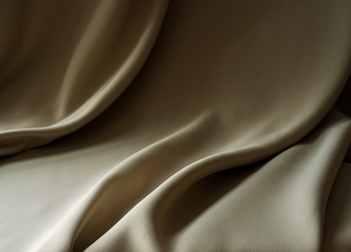Close-up of draped satin fabric in a warm taupe-beige color, showing soft folds and a smooth lustrous texture.