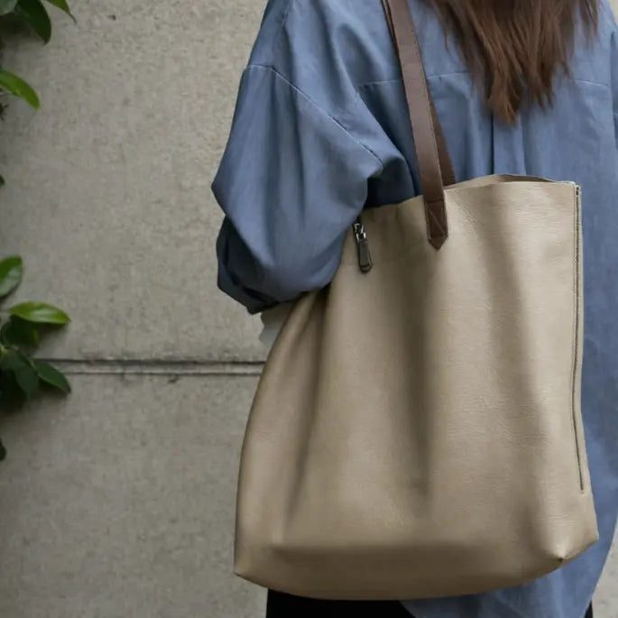 Person holding a beige tote bag with a brown strap against a neutral background
