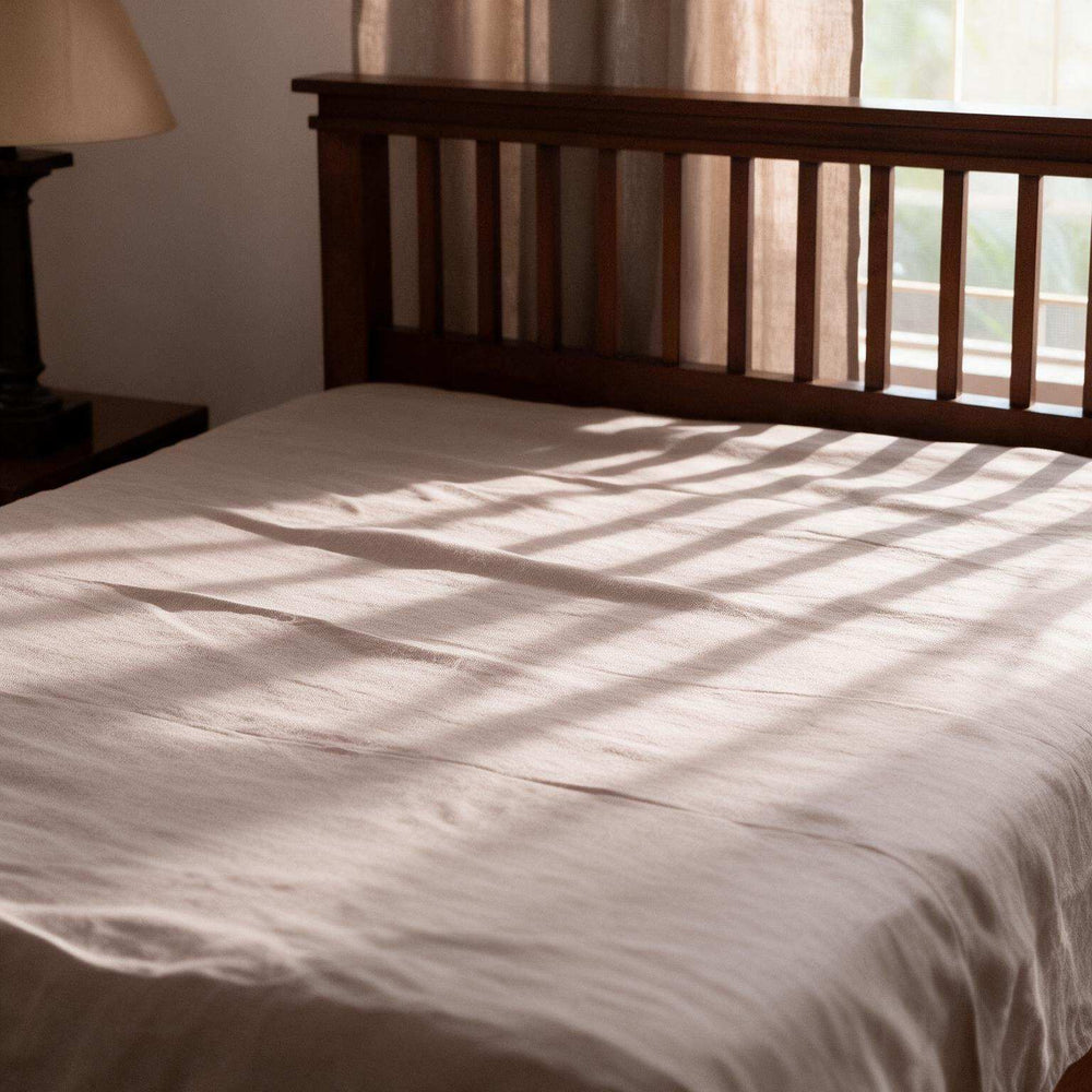 Taupe linen flat sheet set on a dark wood slatted bed with two matching pillowcases, classic table lamp, red clock on the bedside table, and warm morning light through sheer curtains