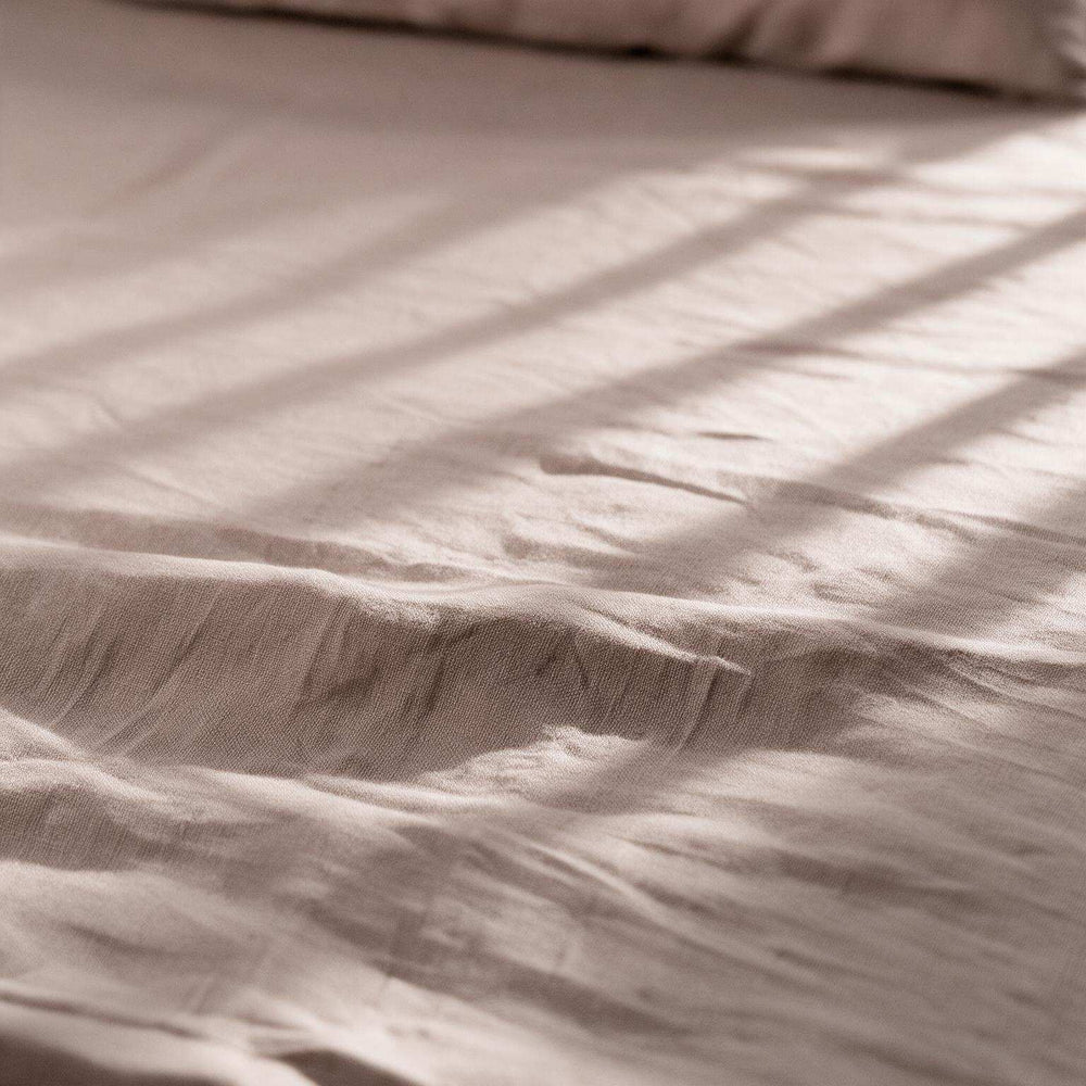 Two Taupe linen pillowcases on a matching flat sheet with dramatic window light casting diagonal stripe shadows across the warm greige stonewashed linen