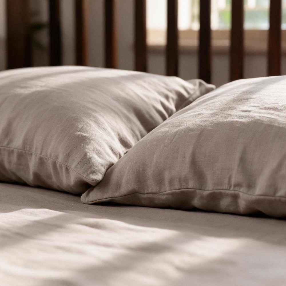 Close-up of two Taupe linen pillowcases on a matching flat sheet with a dark wood headboard and soft dappled light, highlighting the relaxed greige linen drape