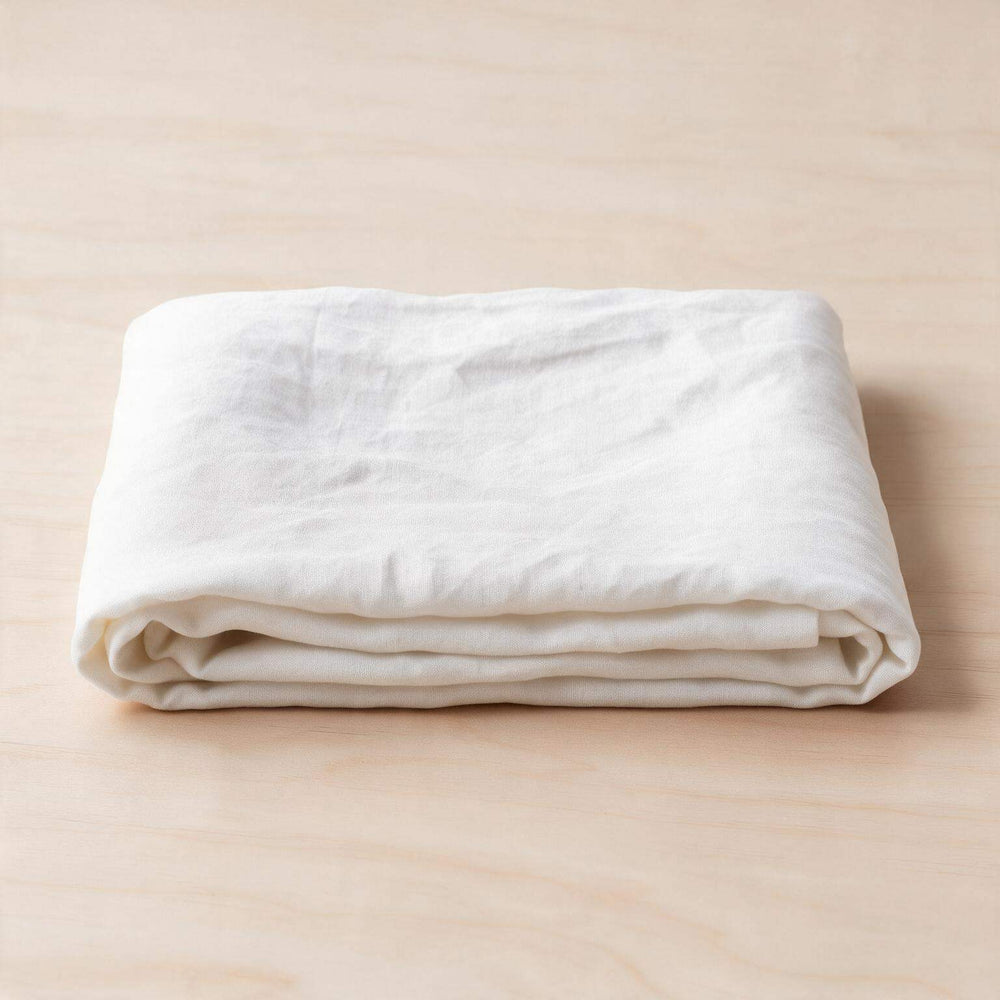 White linen flat sheet set styled on a full bed with two matching pillowcases, light grey upholstered headboard, ceramic lamp, and eucalyptus stems in a bright minimal bedroom
