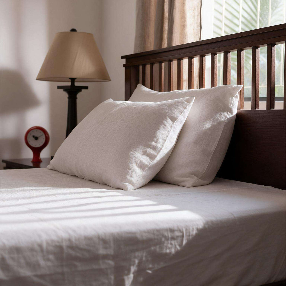 White linen flat sheet set styled on a full bed with two matching pillowcases, light grey upholstered headboard, ceramic lamp, and eucalyptus stems in a bright minimal bedroom