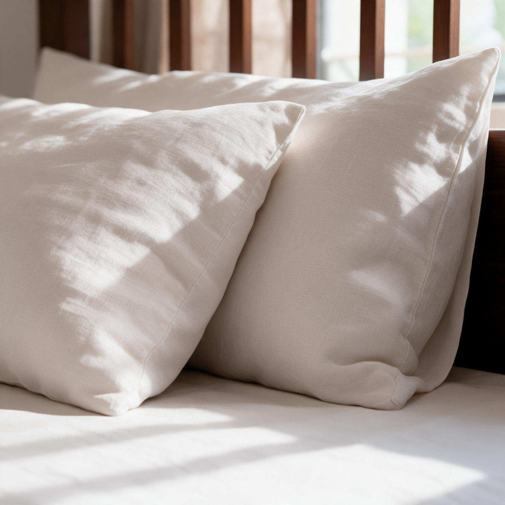 White linen flat sheet set on a bed with oak headboard and matching pillowcases, styled beside a round wooden side table with a potted plant, coffee cup, and book in a bright airy bedroom